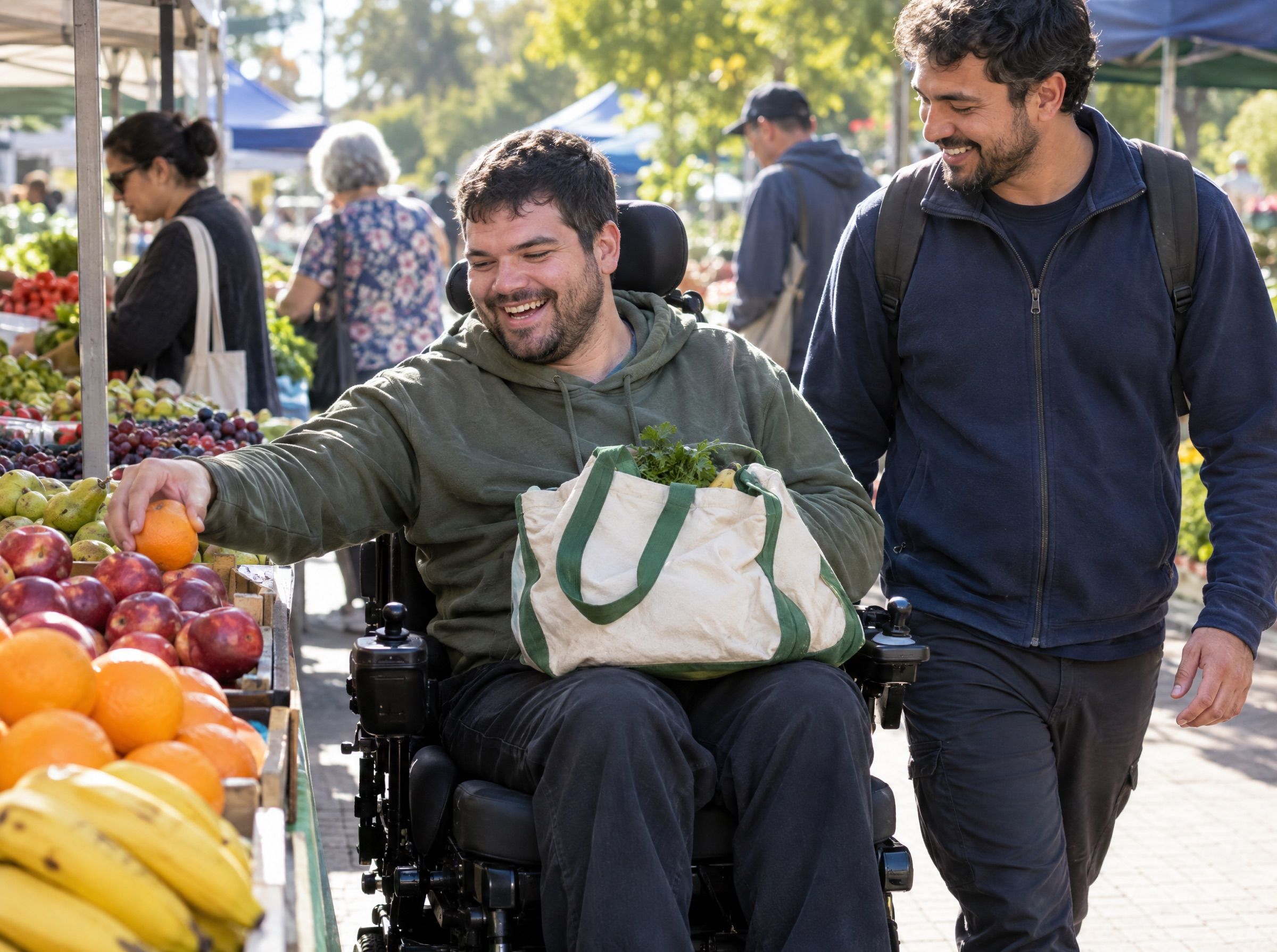 A man in a power wheelchair choosing fruit at a local market, laughing with a support worker beside him.