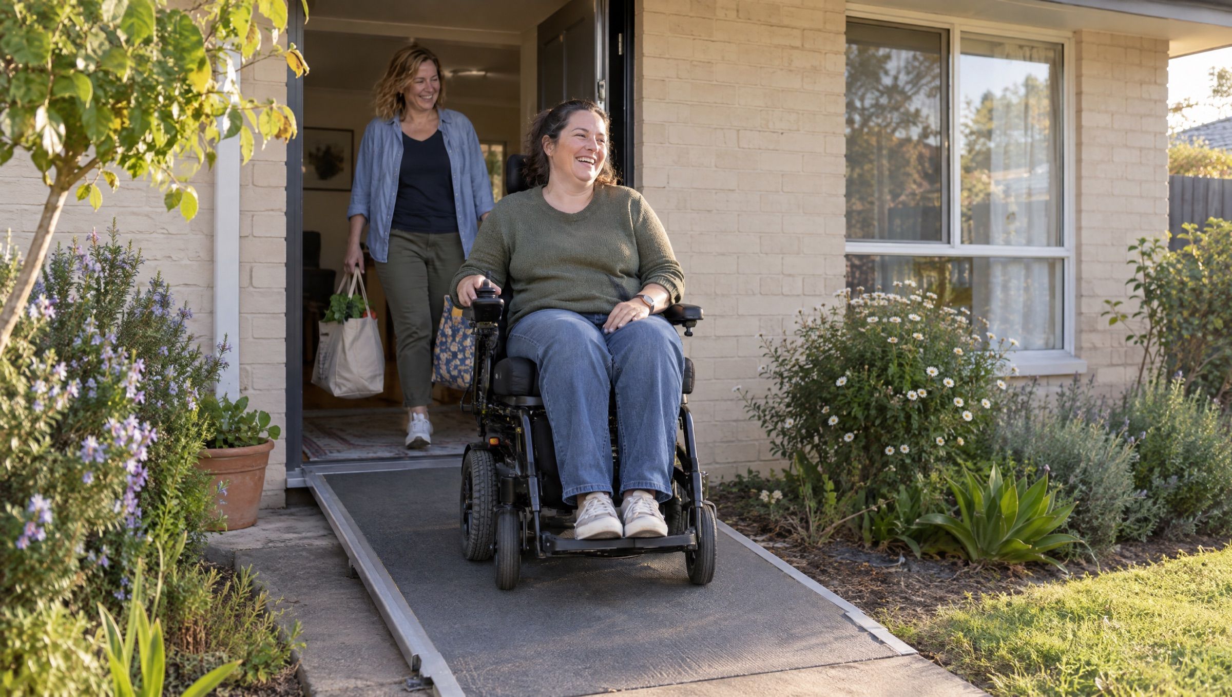 A woman in a power wheelchair coming down the ramp from her front door, with a friend carrying shopping behind her.